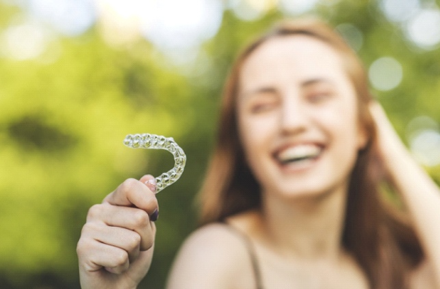 Woman smiling while holding up Invisalign aligner